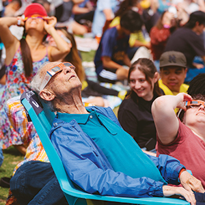 An older man wearing solar eclipse glasses lies back in a chair and looks up at the sun.