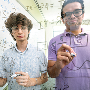 Two young men write mathematical equations on a window.